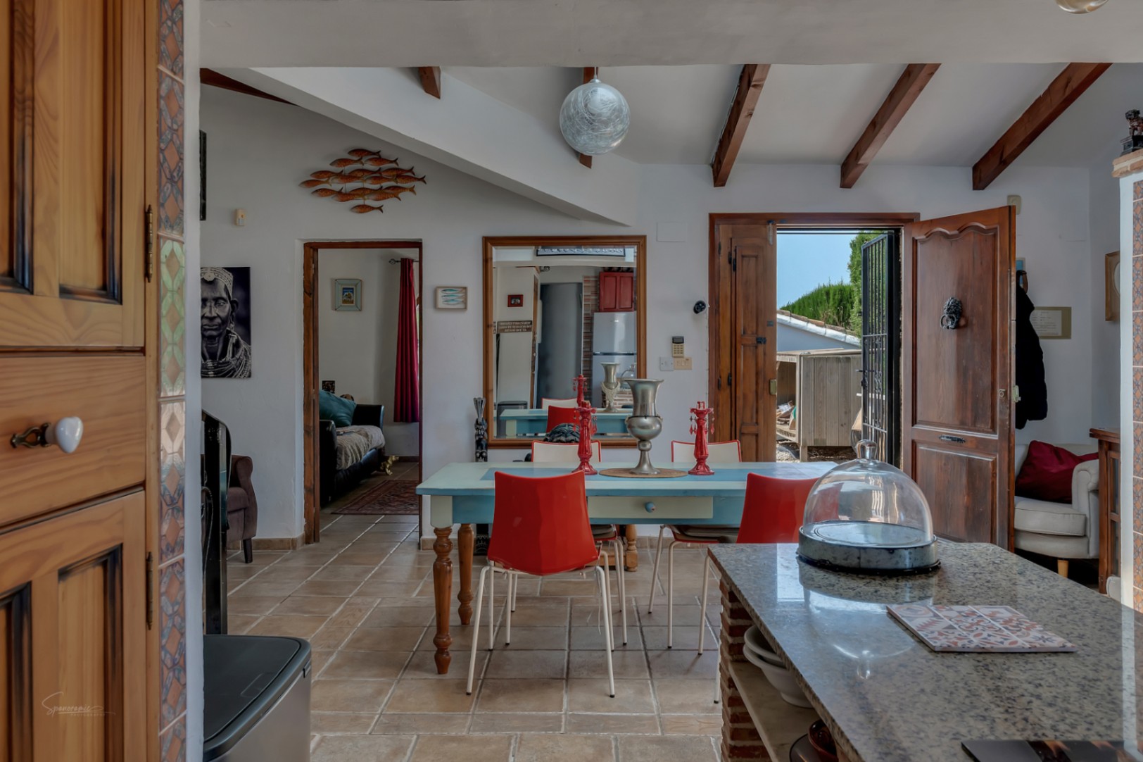 Salle à manger rustique avec table bleue, chaises rouges, sol carrelé et poutres apparentes.
