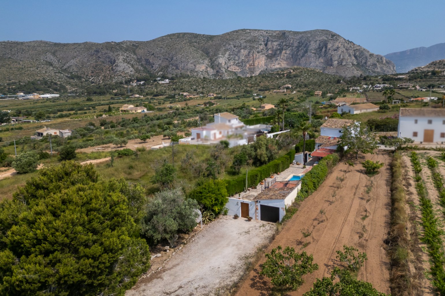 Vue aérienne de la propriété rurale avec allée en terre, garage et petite piscine. Des champs cultivés et des montagnes rocheuses encadrent le paysage de Teulada.