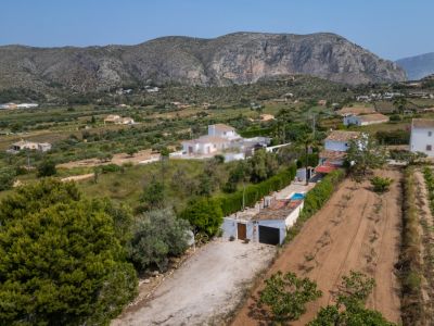 Vue aérienne de la propriété rurale avec allée en terre, garage et petite piscine. Des champs cultivés et des montagnes rocheuses encadrent le paysage de Teulada.