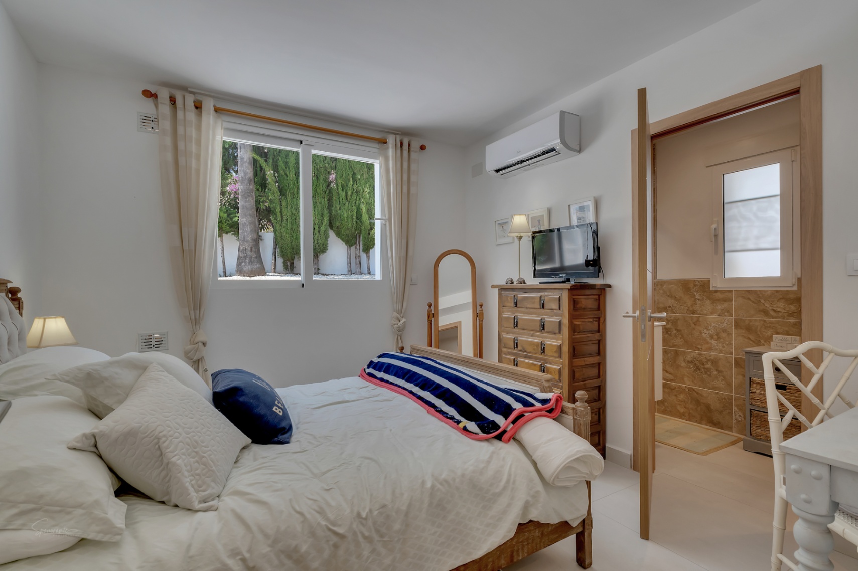 Chambre lumineuse avec lit en bois, commode rustique et salle de bain attenante carrelée dans les tons terre.