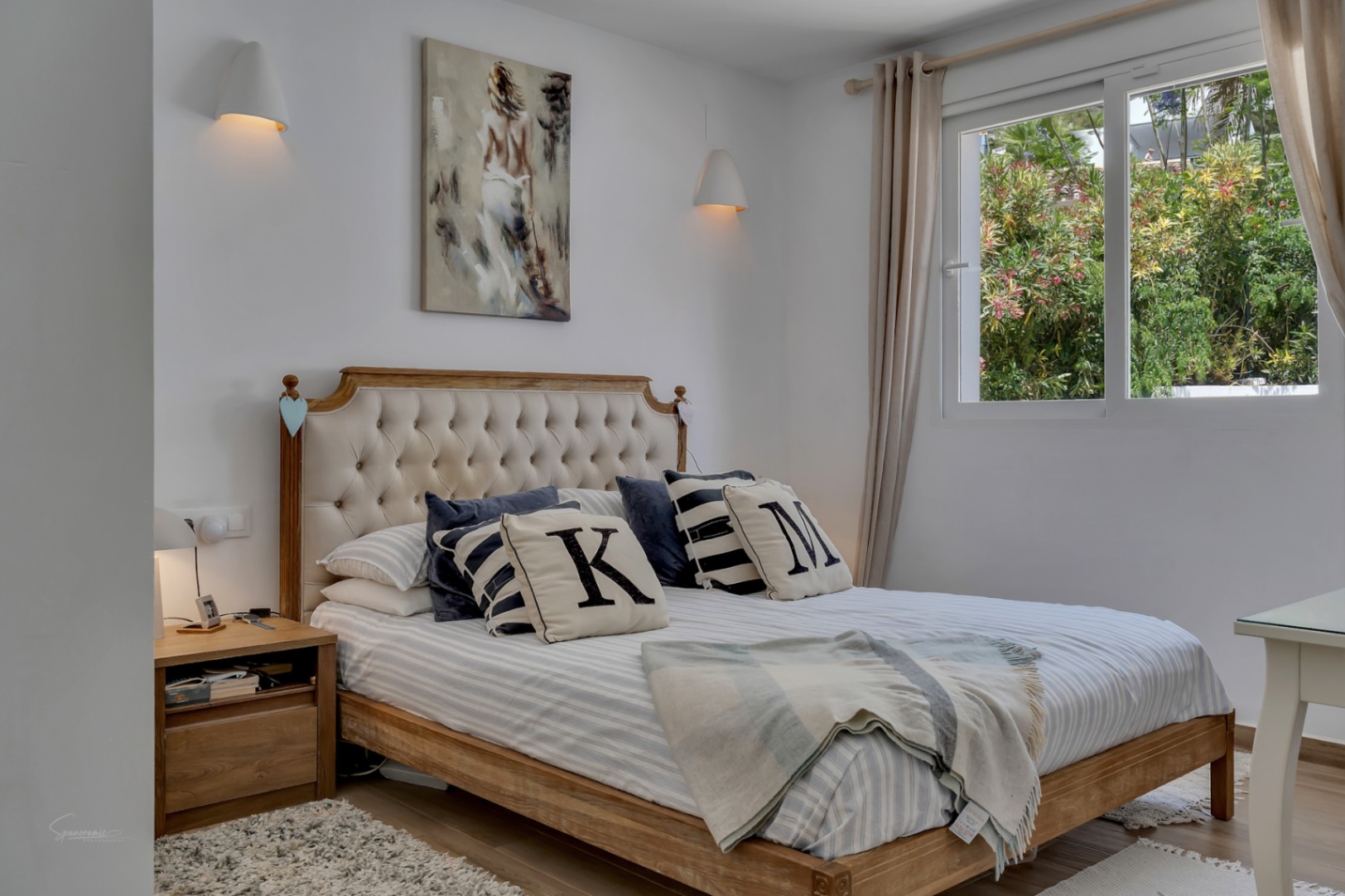 Chambre lumineuse avec lit en bois et tête de lit rembourrée. Décor neutre et lumière naturelle.