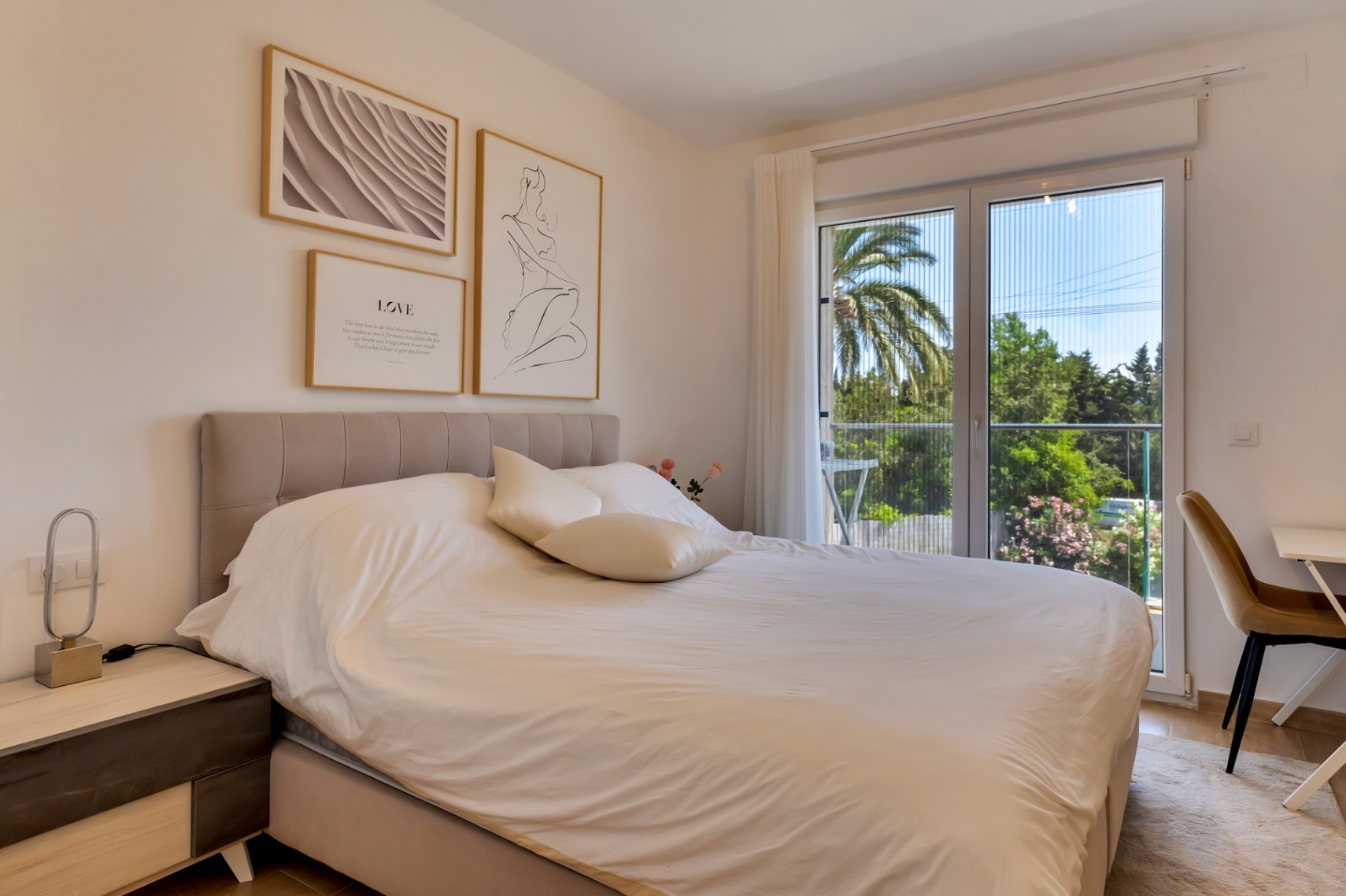 Chambre lumineuse avec lit double, tête de lit rembourrée et coin bureau. Les baies vitrées avec moustiquaire s'ouvrent sur un balcon privé offrant une vue sur la verdure.