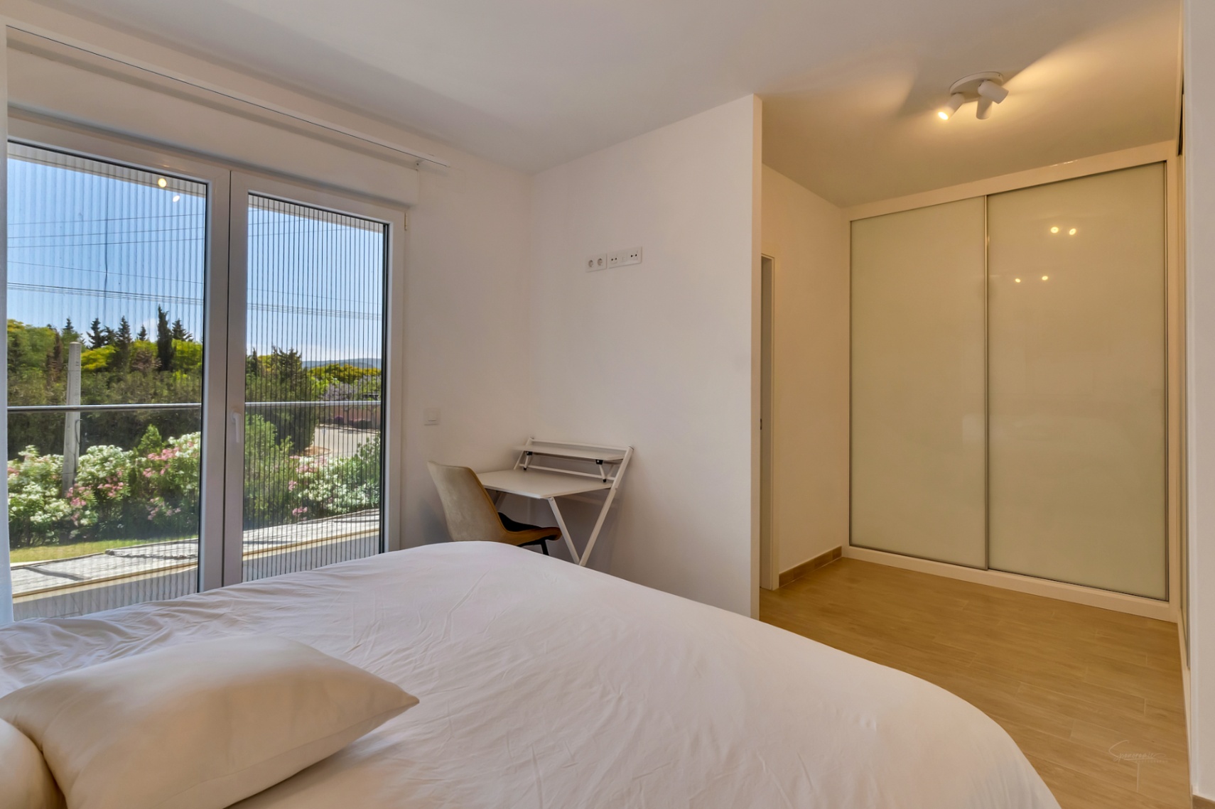 Chambre lumineuse avec placard intégré aux portes coulissantes blanches, coin bureau et accès direct au balcon par une baie vitrée équipée d'une moustiquaire.