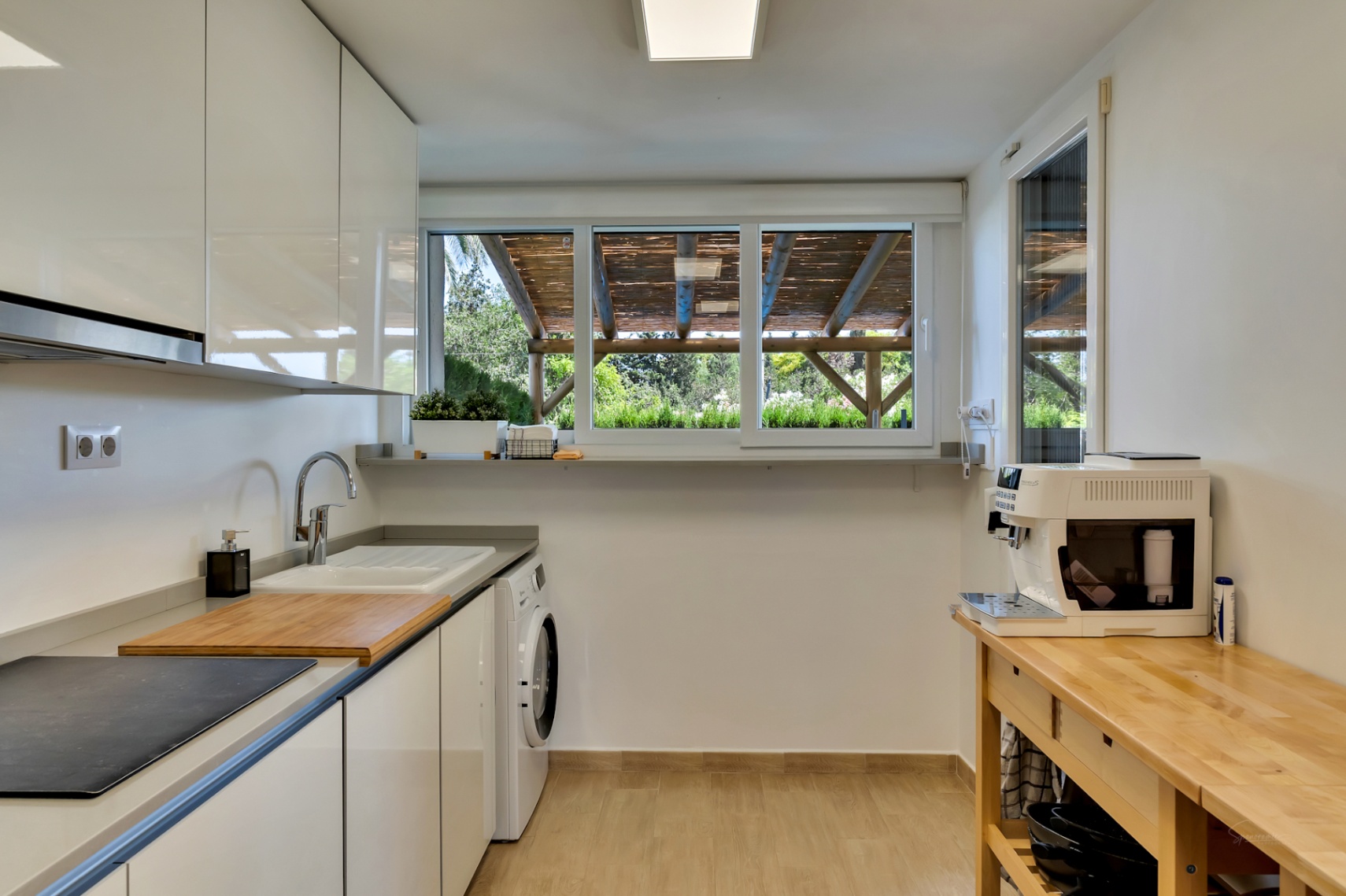 Cuisine moderne avec placards blancs brillants, espace buanderie intégré et une large fenêtre donnant sur la pergola en bois du patio privé.