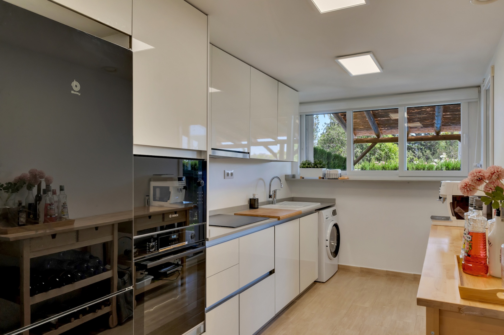 Cuisine moderne avec placards blancs laqués, électroménagers intégrés et grande fenêtre offrant une vue sur le patio privé et sa pergola.