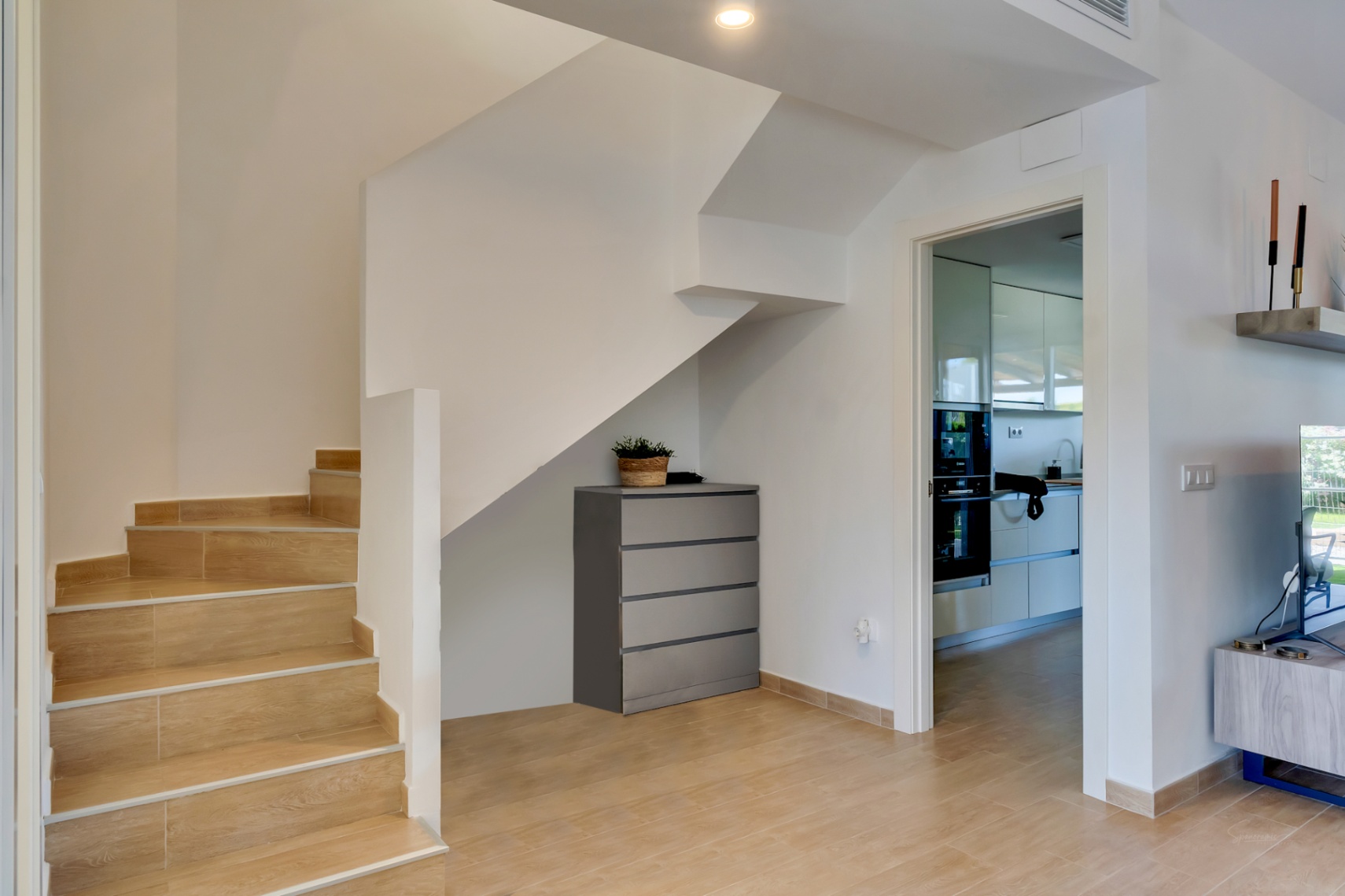 Entrée avec escalier en grès cérame aspect bois, espace de rangement sous l'escalier et vue sur la cuisine moderne équipée.