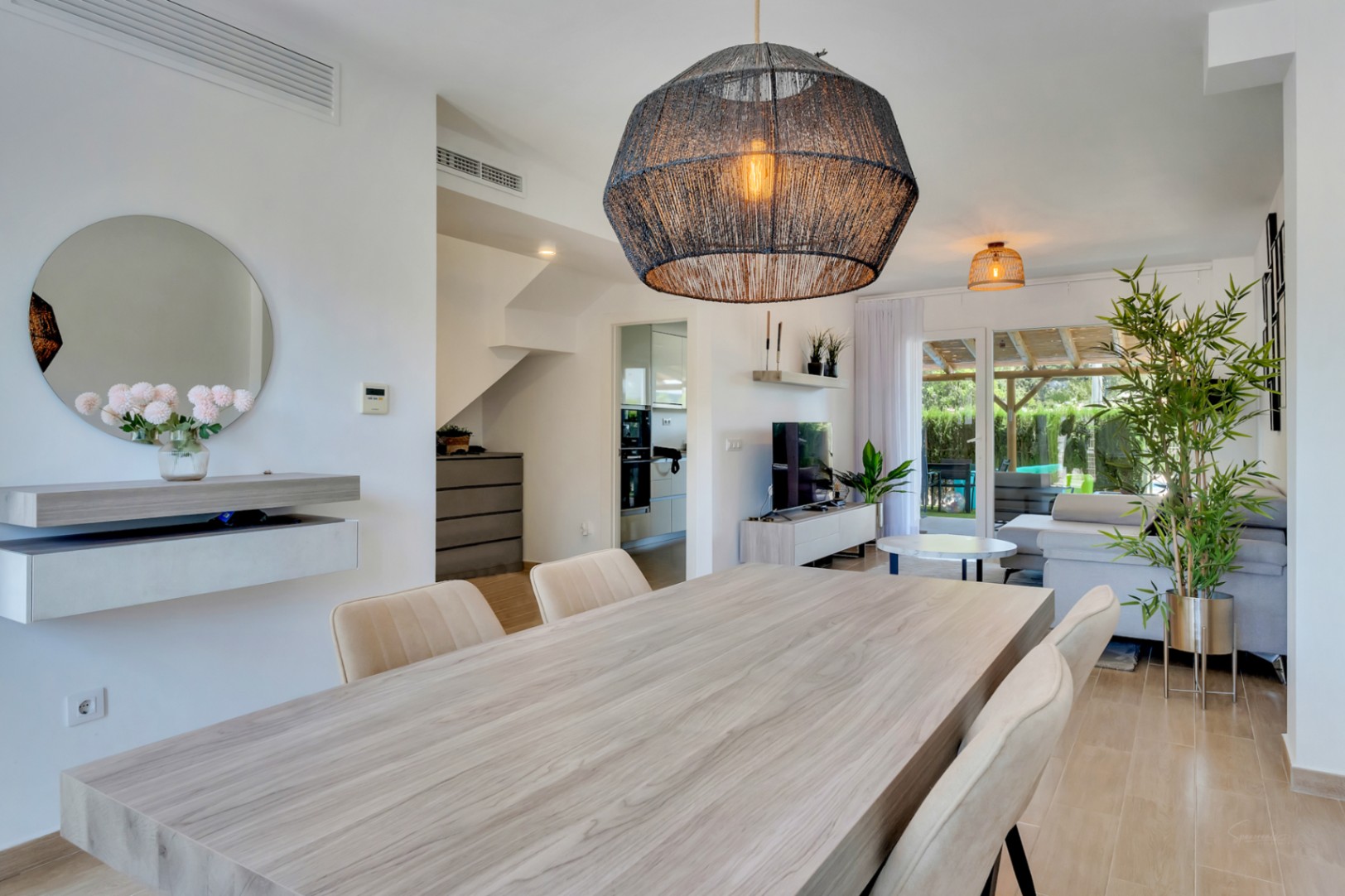 Espace de vie ouvert comprenant une salle à manger avec table en bois, un luminaire en fibres et un accès direct vers la terrasse ombragée.