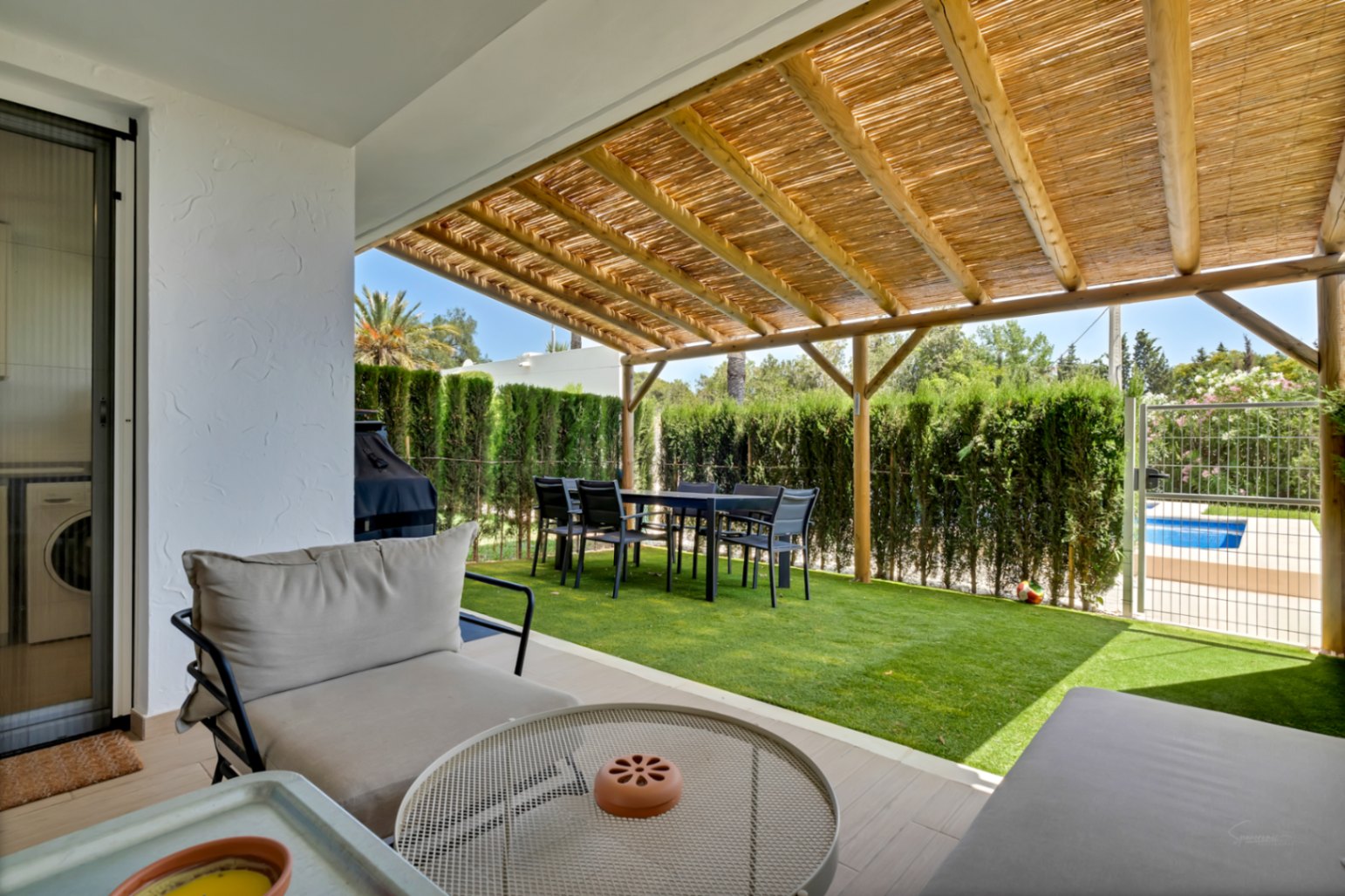 Patio privé avec pergola en bois et canisse, coin repas extérieur et gazon artificiel. Un portillon permet un accès direct à la piscine communautaire et aux jardins.