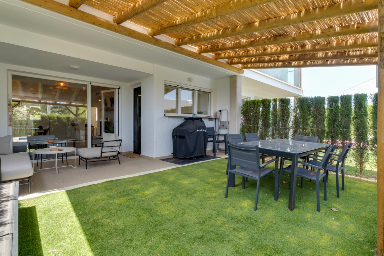 Patio privé avec pergola en bois et canisse, coin repas extérieur et gazon synthétique, relié au salon par de larges baies vitrées coulissantes.