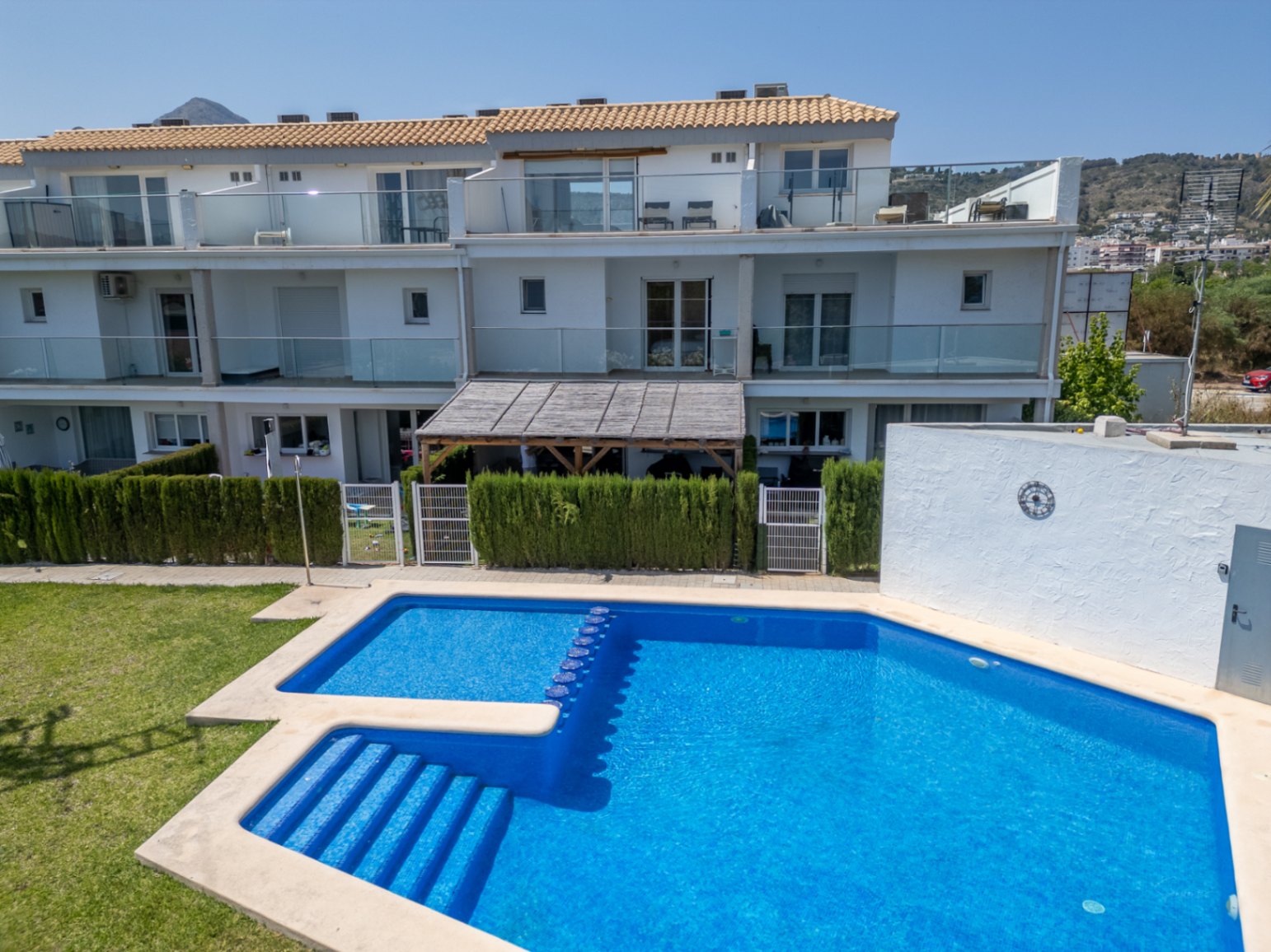 Piscine communautaire aux carreaux bleus avec escaliers intégrés, située devant la façade moderne aux balustrades en verre et patios privés bordés de haies.