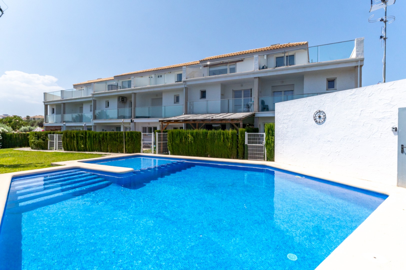Piscine communautaire en mosaïque bleue bordée par la façade moderne des maisons, avec balustrades en verre et jardins privés délimités par des haies verdoyantes.