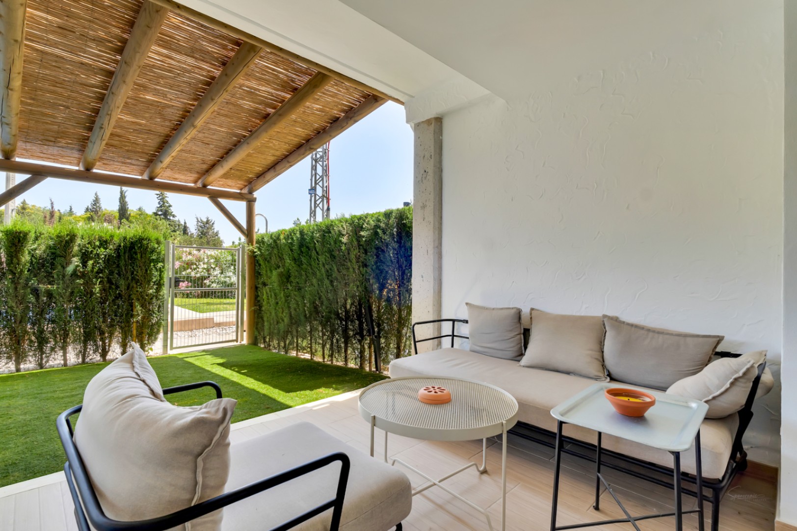 Salon d'été couvert sous une pergola en bois et canisse, doté de mobilier contemporain et ouvrant sur un jardin privé avec gazon synthétique et haies de cyprès.