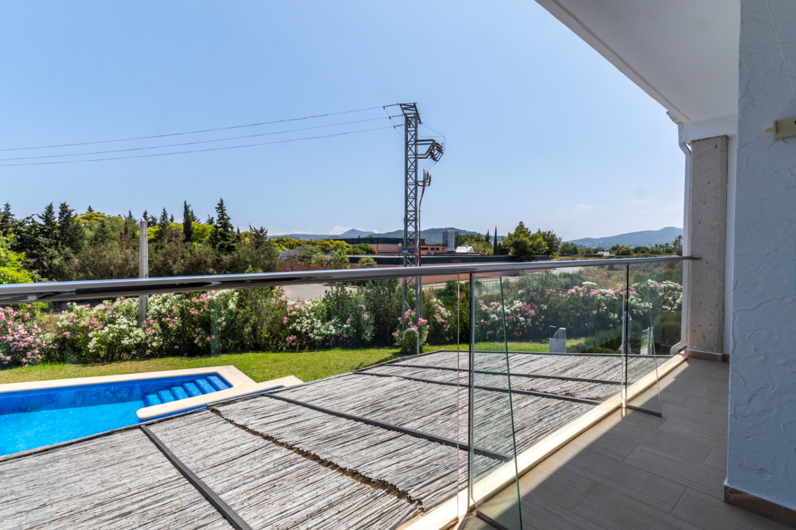 Terrasse à l'étage avec garde-corps en verre offrant une vue sur le patio privé ombragé par une canisse, la piscine et les collines environnantes.