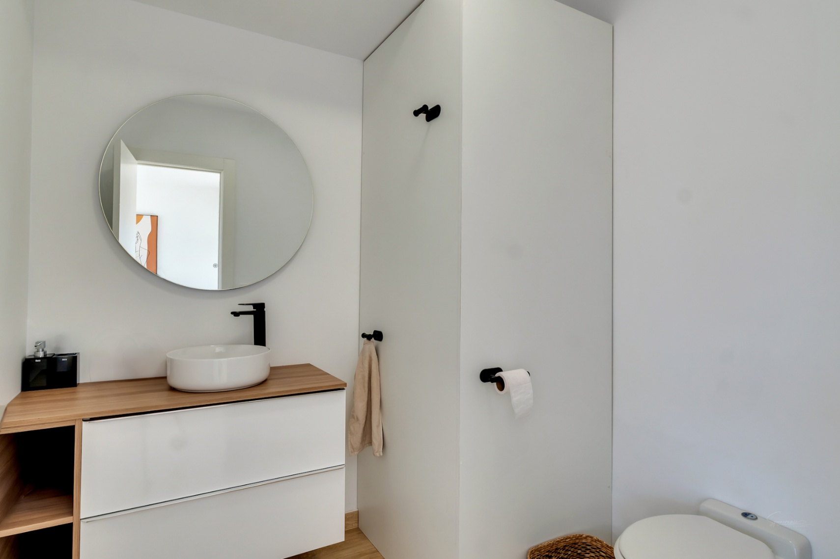 Toilettes modernes avec vasque à poser sur plan en bois, robinetterie noire mate et miroir circulaire. Le meuble suspendu blanc souligne un design épuré et contemporain.