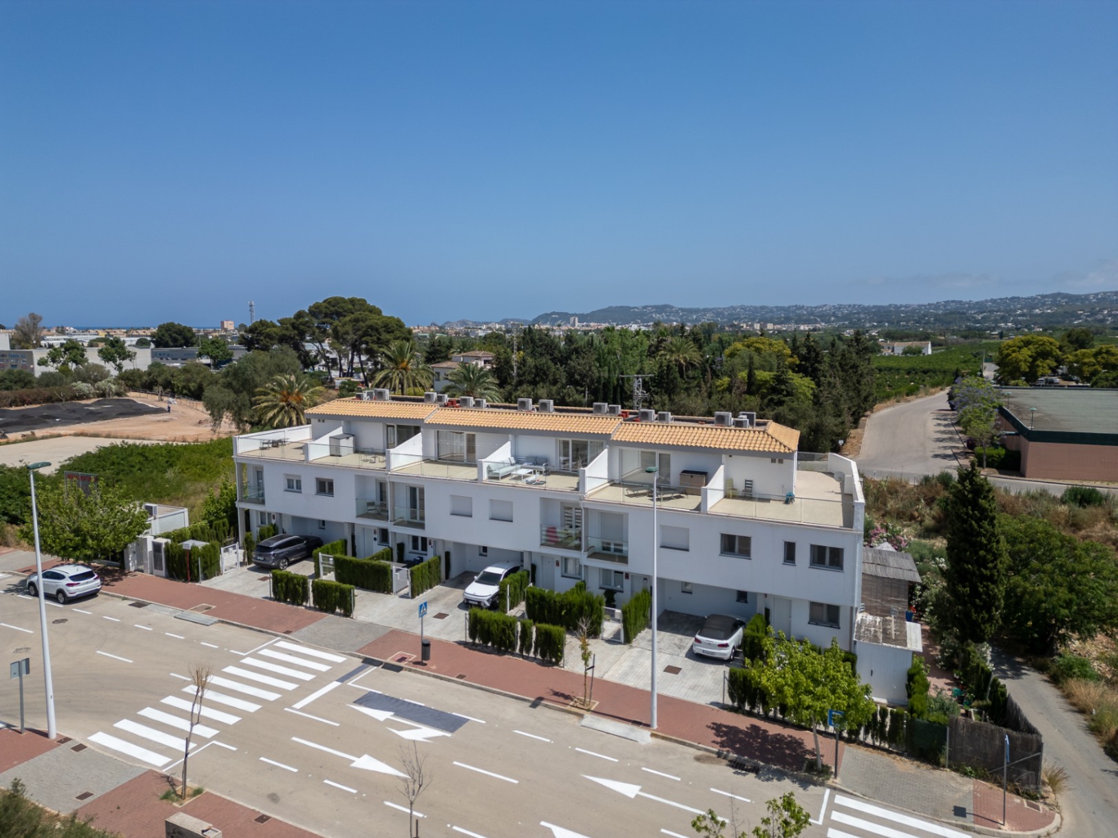 Vue extérieure de l'ensemble de maisons mitoyennes modernes aux façades blanches, avec terrasses privées, garde-corps en verre et places de parking individuelles.