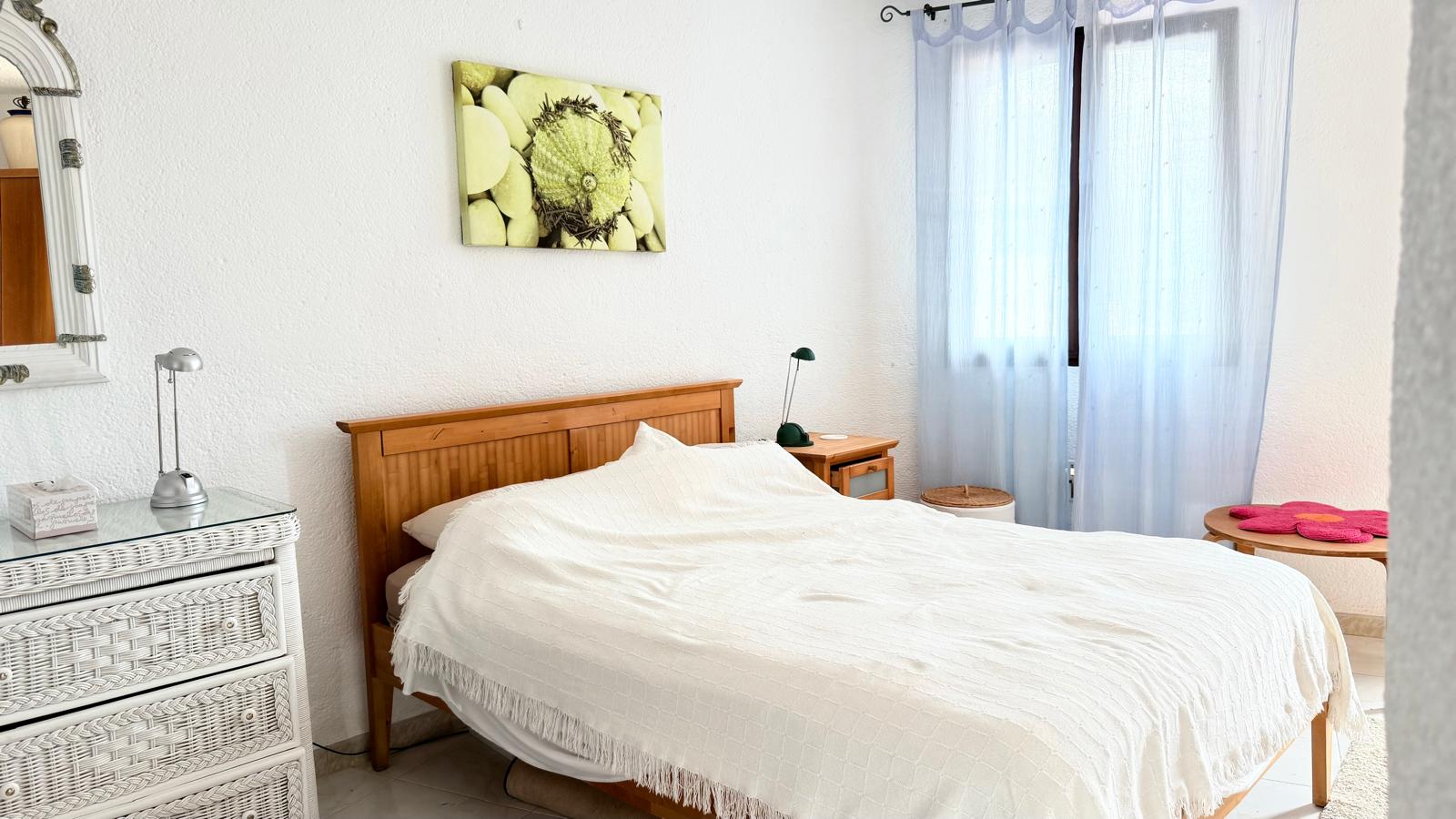 Chambre avec cadre de lit en bois, commode en osier blanc et murs texturés. La pièce bénéficie d'une lumière naturelle filtrée par des rideaux bleus translucides.