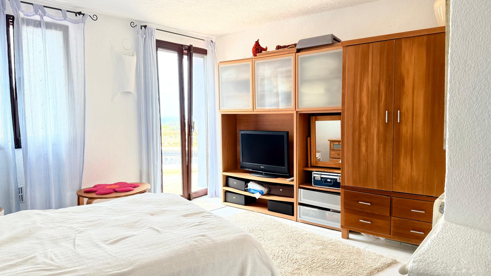 Chambre lumineuse avec placards en bois intégrés, coin TV et accès direct à une terrasse privée offrant une vue dégagée sur le paysage littoral.