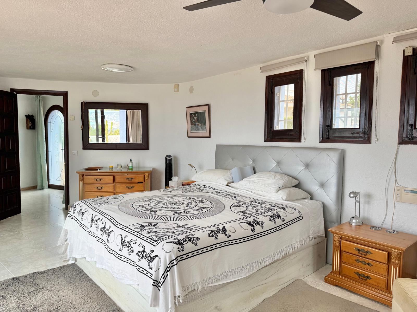 Chambre principale avec murs incurvés, mobilier en bois classique et fenêtres à cadres sombres. La pièce dispose d'un ventilateur de plafond et d'un sol carrelé clair.