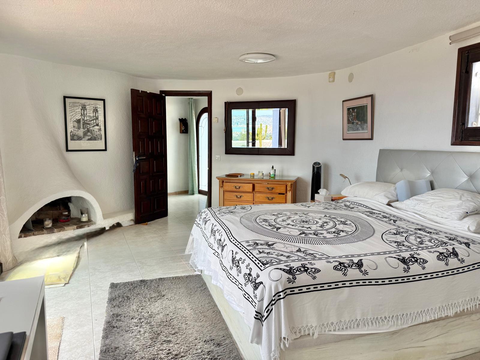 Chambre principale de style ibizane avec cheminée d'angle intégrée, murs blancs texturés et carrelage clair offrant une atmosphère lumineuse et authentique.