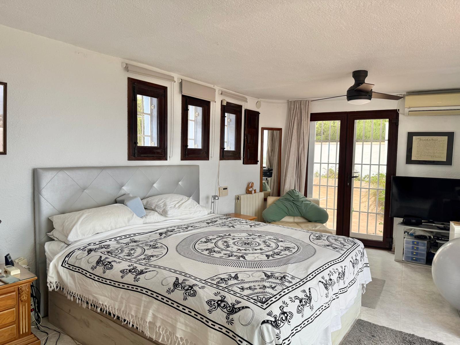 Chambre principale lumineuse avec menuiseries en bois sombre, ventilateur de plafond et accès direct à la terrasse par des doubles portes vitrées.
