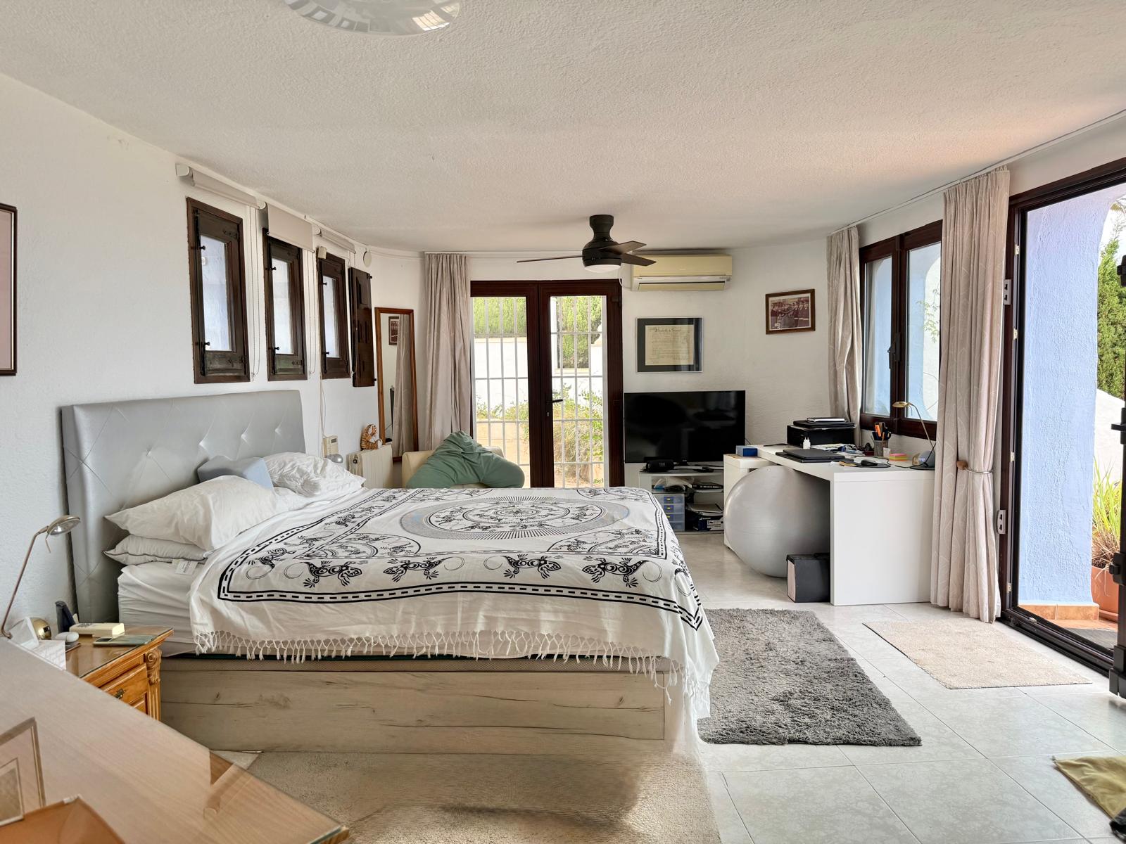 Chambre principale spacieuse avec accès direct à la terrasse, coin bureau, ventilateur de plafond, climatisation et menuiseries en bois sombre.