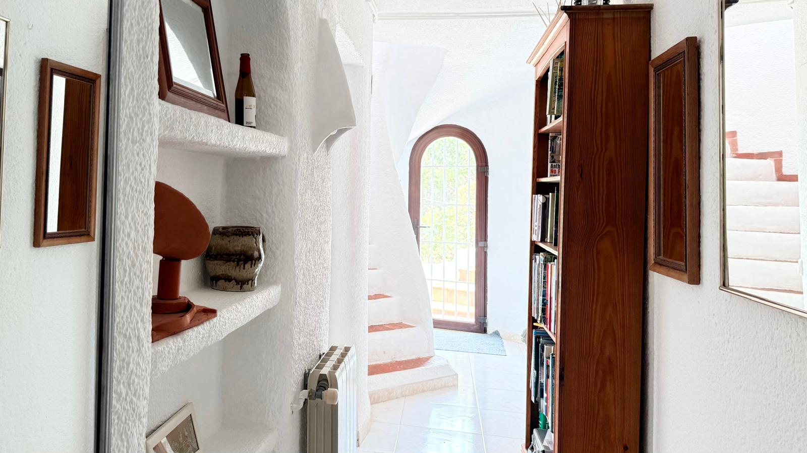 Couloir de style ibizane avec murs texturés blancs, étagères encastrées en maçonnerie et un escalier courbe aux nez de marches en terre cuite baigné de lumière naturelle.