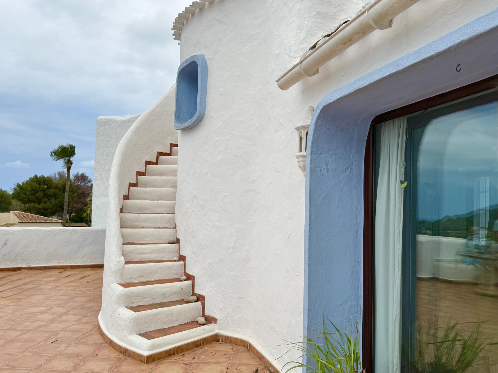 Escalier extérieur incurvé de style ibizenc avec marches en terre cuite, intégré à la façade blanche texturée et encadrements de fenêtres bleus.