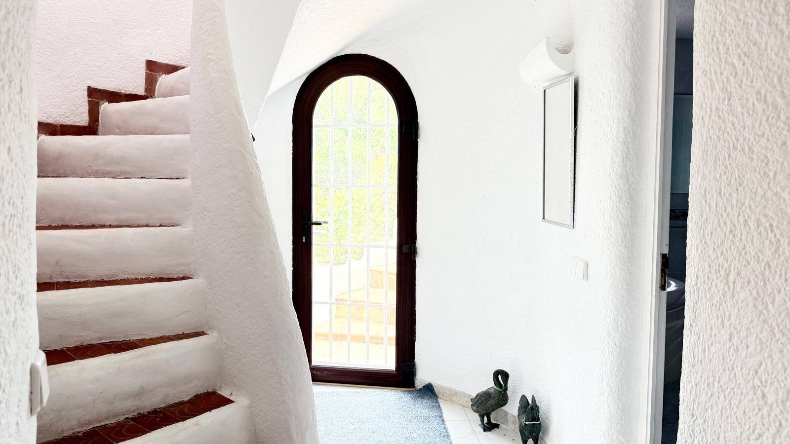 Escalier intérieur courbe de style ibizane avec des marches en terre cuite et des murs blancs texturés, situé à côté d'une porte cintrée vitrée.