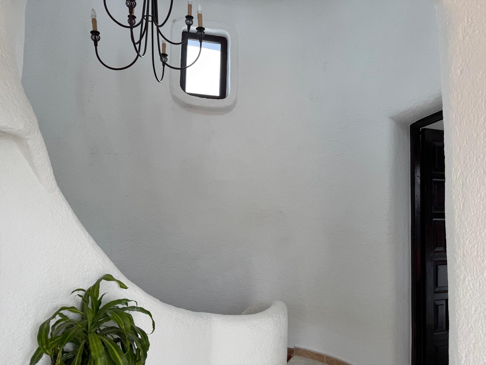 Escalier intérieur courbe de style ibizien avec des murs en plâtre blanc texturé, une petite fenêtre carrée et un lustre élégant en fer forgé.