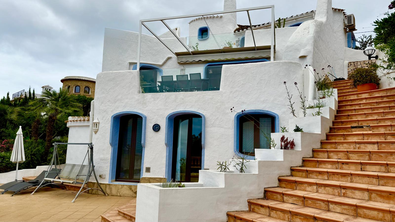 Extérieur de style Ibiza avec murs blancs et encadrements bleus. Des escaliers en terre cuite relient les différents niveaux et terrasses équipées de garde-corps en verre.