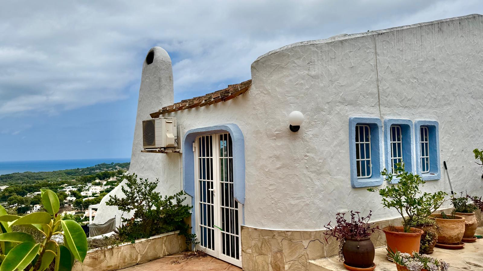 Façade de style Ibiza avec murs blancs arrondis, menuiseries bleues et vue panoramique sur la côte méditerranéenne depuis la terrasse.