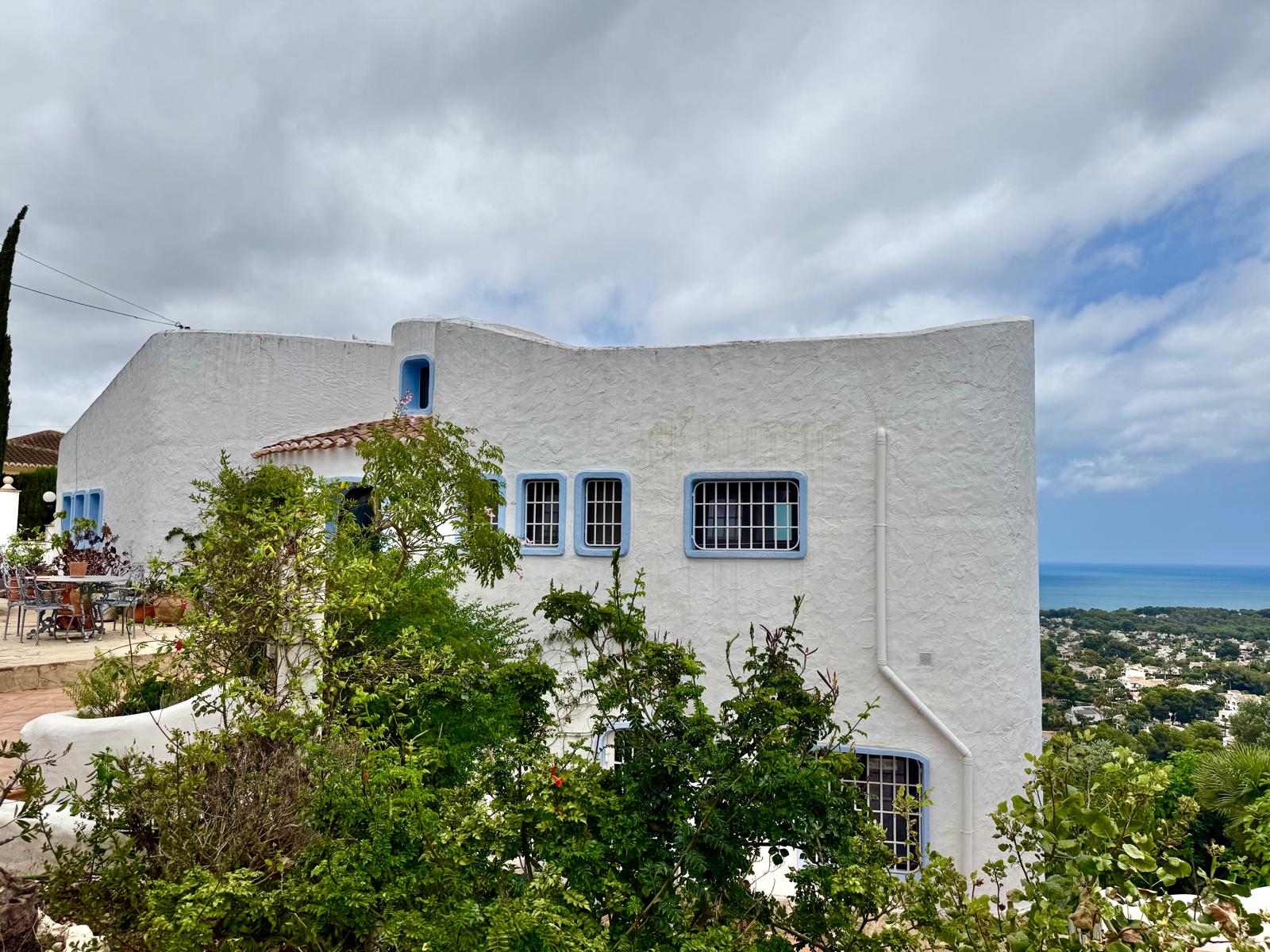 Façade de style ibizien aux murs blancs incurvés et menuiseries bleues, offrant une vue imprenable sur le littoral de Jávea depuis sa position dominante.