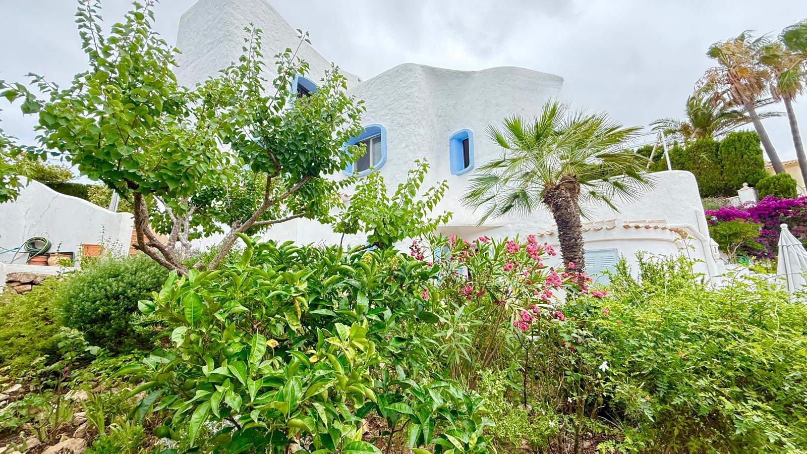Façade de style ibizien aux murs blancs texturés et fenêtres bleues, entourée d'un jardin méditerranéen luxuriant avec des palmiers et des lauriers-roses.
