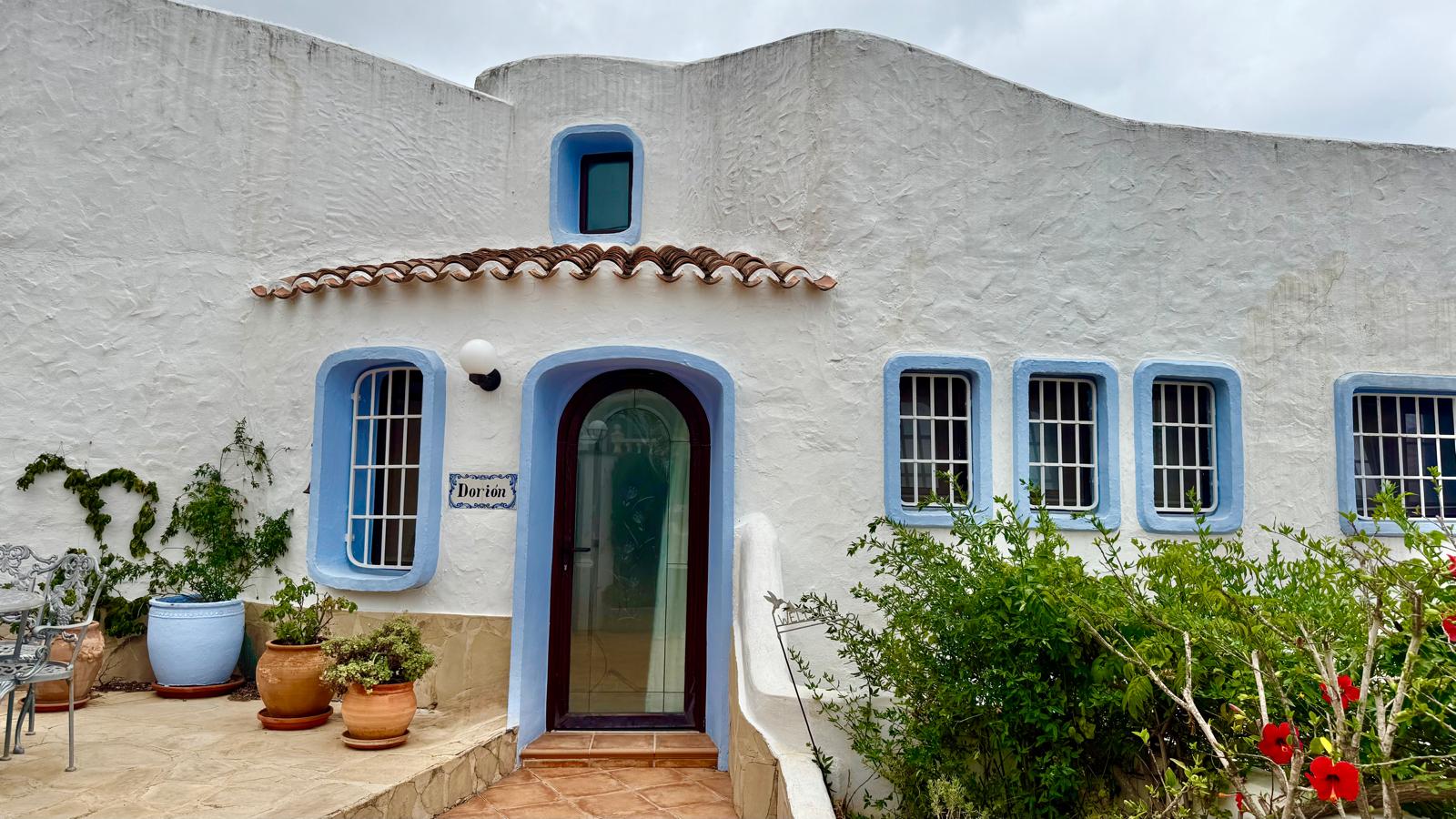 Façade de style ibizien aux murs blancs texturés, fenêtres soulignées de bleu et porte voûtée. Des pots en terre cuite et une végétation méditerranéenne ornent l'entrée.
