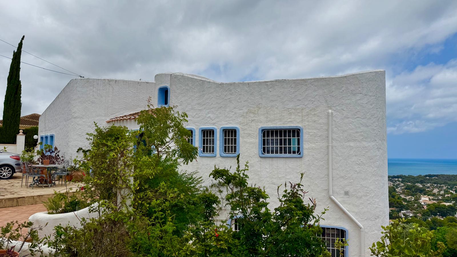Façade de style ibizien aux murs blancs texturés, menuiseries bleues et lignes courbes, offrant une vue panoramique sur la mer depuis sa position dominante.