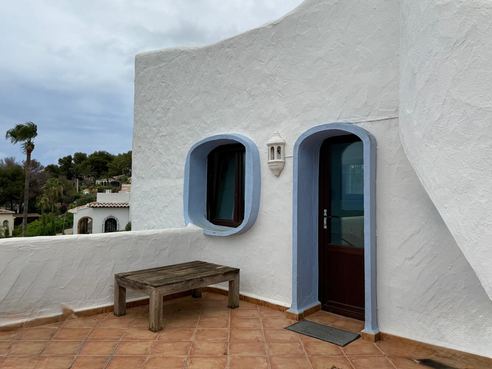 Façade de style ibizien aux murs blancs texturés, menuiseries en bois sombre et encadrements arrondis bleus. La terrasse en terre cuite offre un espace extérieur authentique et épuré.