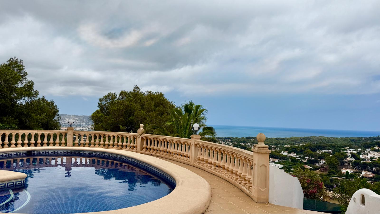 Piscine circulaire avec mosaïque bleue et balustrade classique en pierre, offrant une vue panoramique sur la mer Méditerranée et le paysage de Jávea.