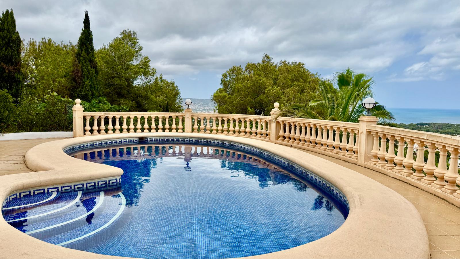Piscine privée aux formes courbes avec mosaïque bleue, entourée d'une terrasse en pierre et d'une balustrade classique offrant une vue panoramique sur la mer.
