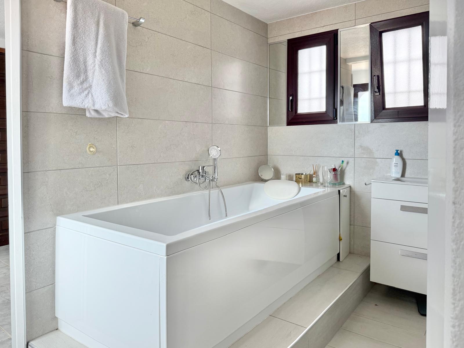 Salle de bain avec baignoire blanche et repose-tête, murs en grès cérame aux tons neutres et meuble vasque blanc à côté de fenêtres aux cadres sombres.