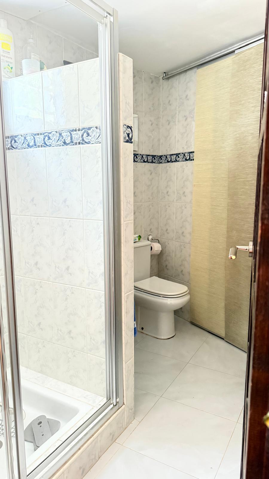 Salle de bain avec cabine de douche vitrée, carrelage blanc orné d'une frise bleue et toilettes blanches. Une porte coulissante texturée apporte une touche d'intimité.