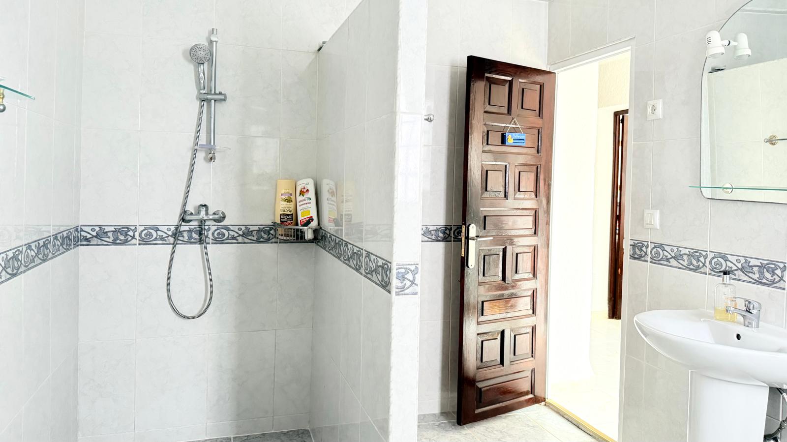 Salle de bain avec douche à l'italienne, carrelage blanc à frise décorative, lavabo sur colonne et porte en bois massif sculpté.