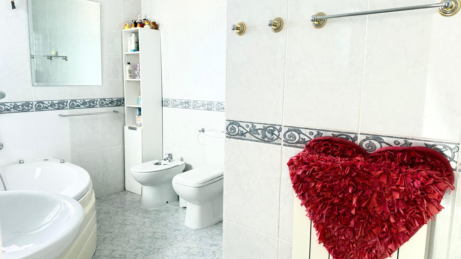 Salle de bain comprenant une baignoire d'angle à remous, un bidet et des toilettes, avec des murs carrelés blancs et une frise décorative.