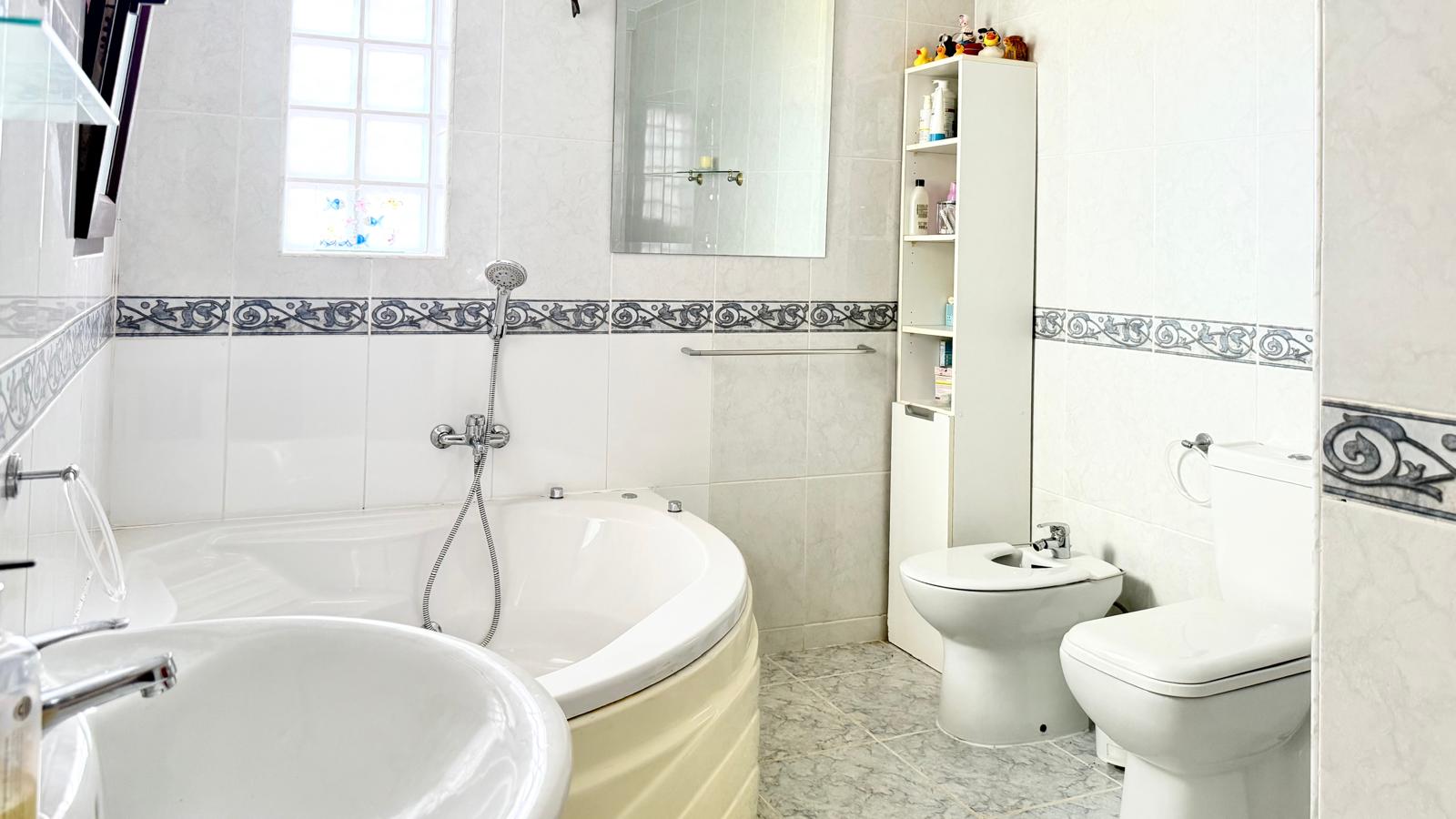 Salle de bains équipée d'une baignoire d'angle avec hydromassage, d'un bidet et de toilettes, avec des murs carrelés blancs et une frise décorative classique.