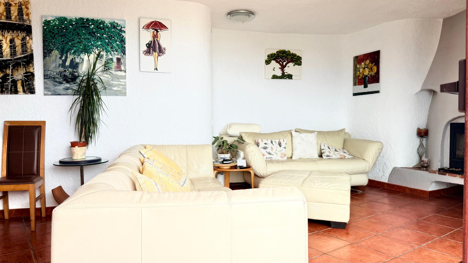 Salon de style ibizenc avec sol en terre cuite, murs blancs texturés et cheminée à bois. L'espace comprend des canapés en cuir crème et une décoration murale.