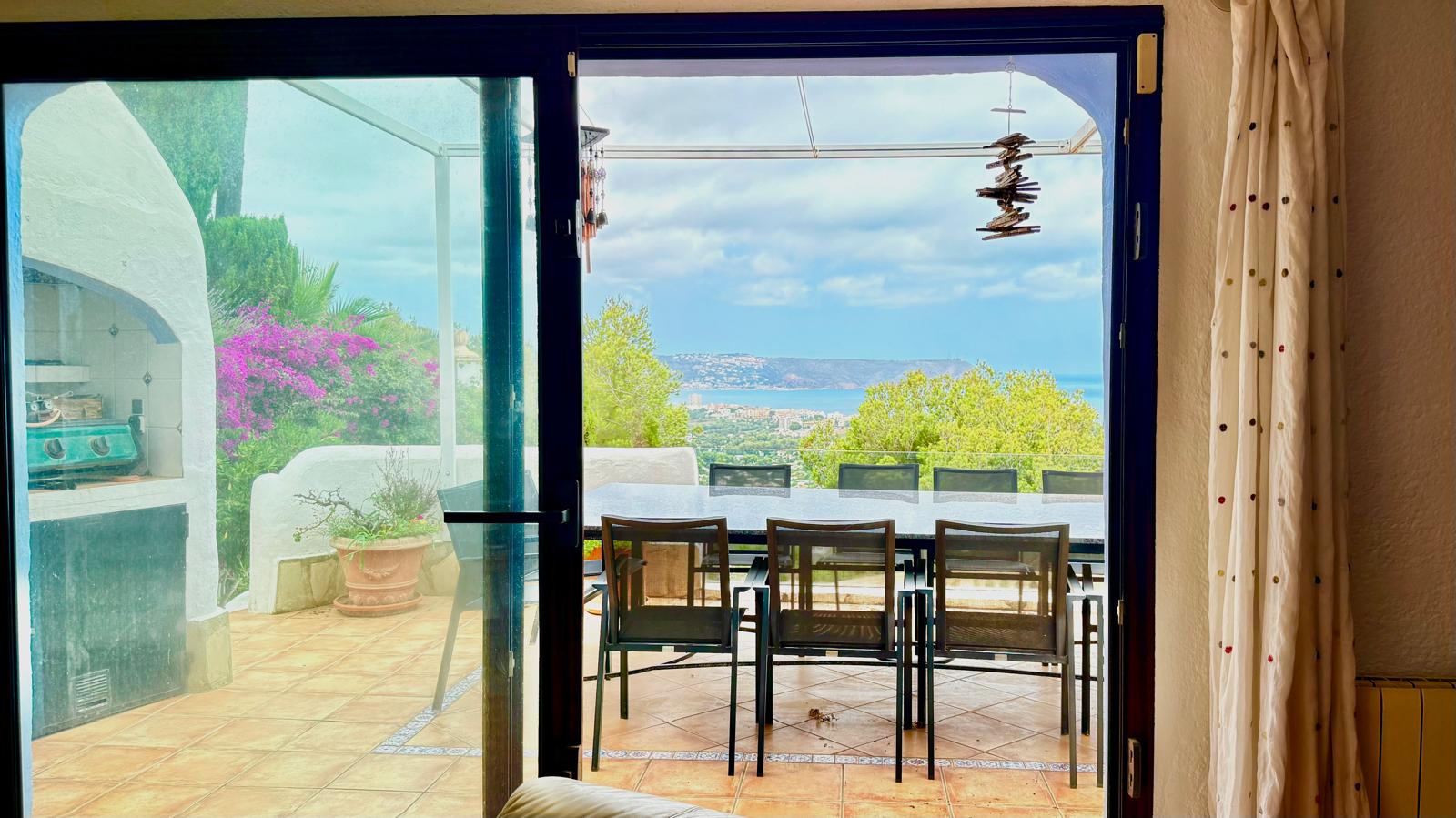 Vue depuis l'intérieur vers la terrasse principale avec espace repas, barbecue maçonné et vue panoramique sur la baie de Jávea et la mer Méditerranée.