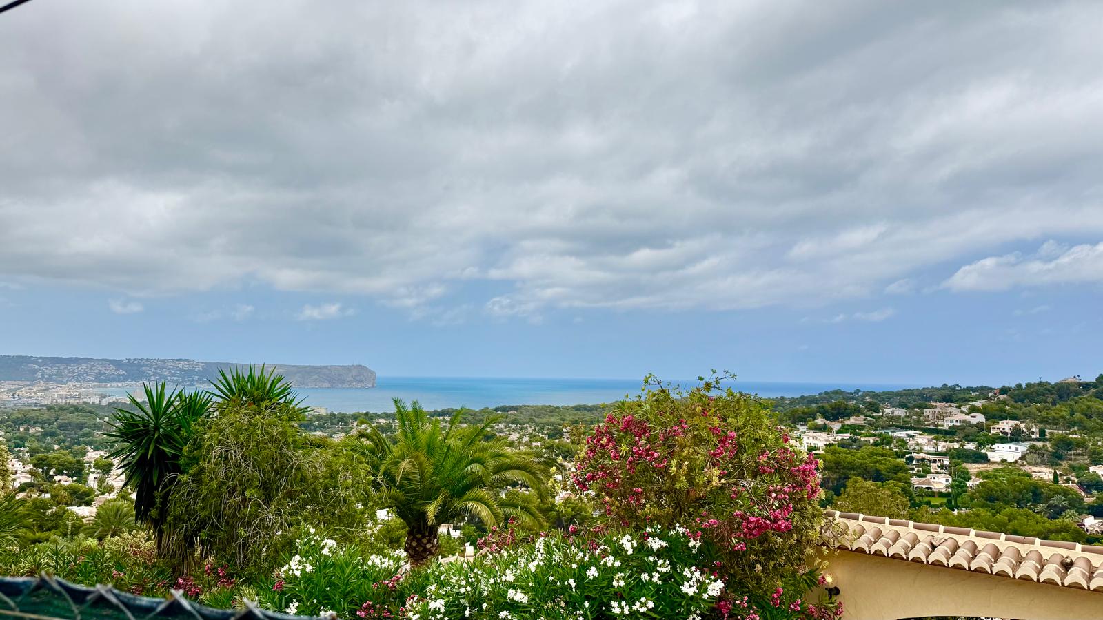 Vue panoramique sur la baie de Jávea et le Cap San Antonio, entourée d'un jardin méditerranéen luxuriant avec des lauriers-roses et des palmiers.
