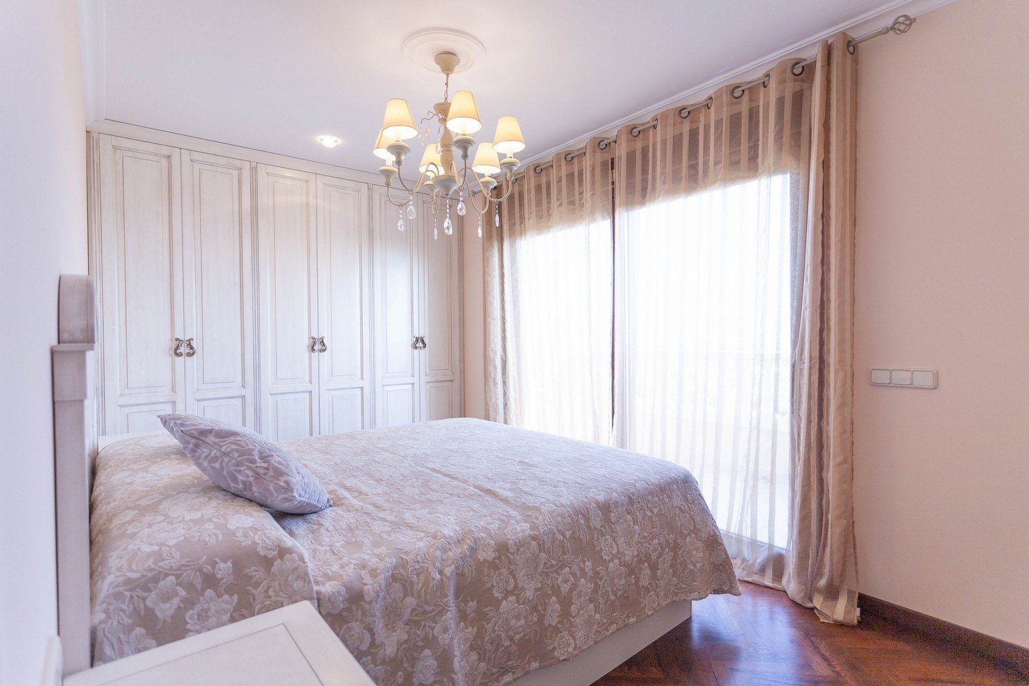 Chambre avec lit double, armoire intégrée blanche, lustre et rideaux beiges.