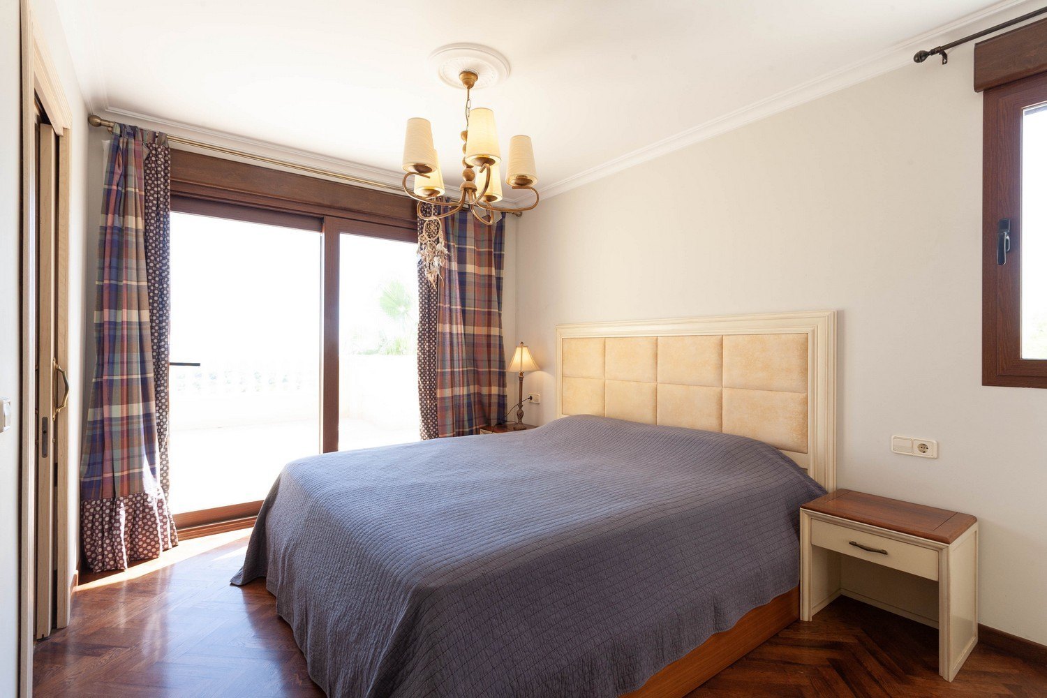 Chambre avec lit double, tête de lit rembourrée et accès à la terrasse. Lustre classique et rideaux à motifs.