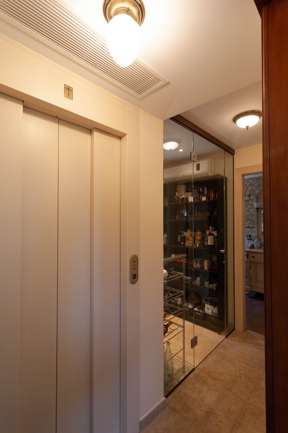 Détail de l'ascenseur intérieur à côté d'un cellier vitré avec des étagères métalliques.