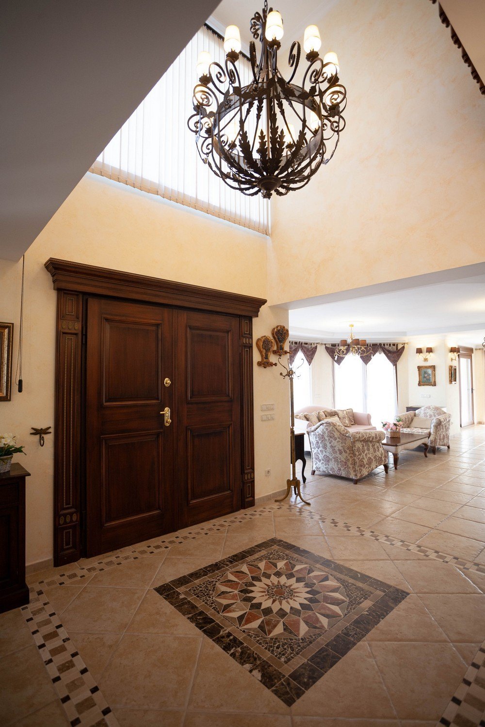 Hall d'entrée avec porte double en bois foncé et lustre orné. Sol carrelé avec motif géométrique.